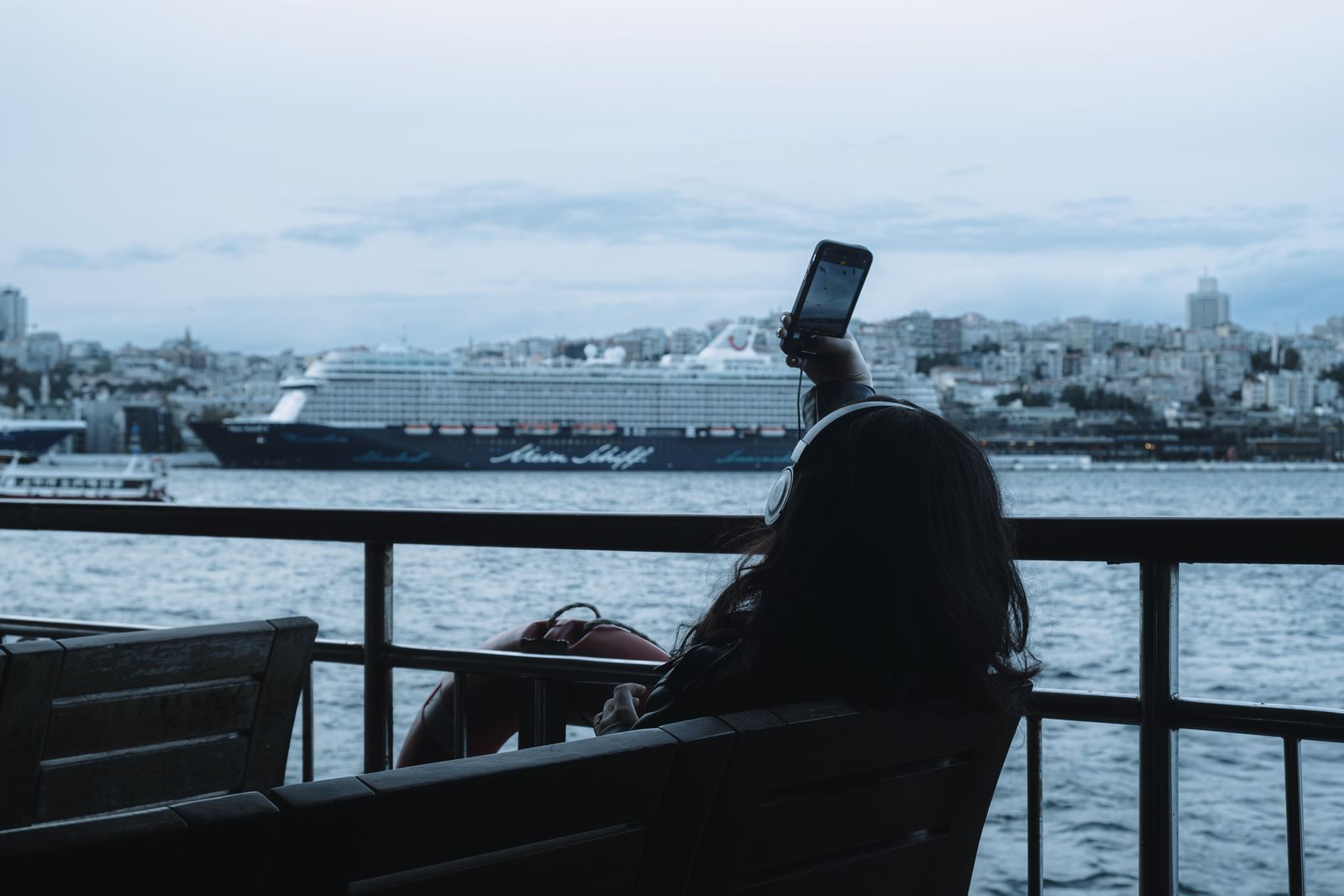 Traveler on a waterfront terrace holding up a phone, with a cruise ship docked beyond, checking cruise fare alerts